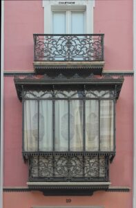 Verzierter Balkon aus Schmiedeeisen vor rosafarbener Fassade mit Fenster und Vorhängen