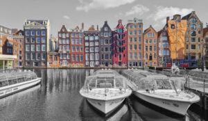 Schwarz-Weiß-Ansicht des Amsterdamer Kanals mit Hausbooten und farbigen, schmalen Giebelhäusern im Hintergrund unter grauem Himmel.