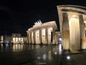 Nächtliche Ansicht des Brandenburger Tors in Berlin, beleuchtet und umrahmt von der Quadriga, mit Passanten im Vordergrund und Gebäuden im Hintergrund.