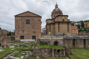 Historische Ruinen des Forum Romanum mit Kirche Santi Luca e Martina in Rom, umgeben von antiken Säulenresten und moderner Bebauung im Hintergrund bei bewölktem Himmel.