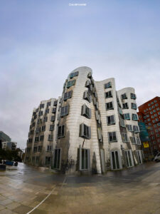 Modernes Gebäude mit geschwungener Metallfassade, unregelmäßig verteilten Fenstern und städtischem Platz bei bewölktem Himmel.