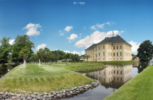 Historisches gelbes Wasserschloss mit weißen Fensterläden, dunklem Schieferdach und Baumallee, reflektiert im ruhigen Wasserspiegel.
