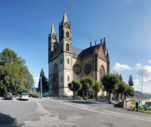 Panorama einer gotischen Kathedrale mit dualen Spitztürmen, großem Rosettenfenster und Wasser- sowie Hügelblick unter blauem Himmel.