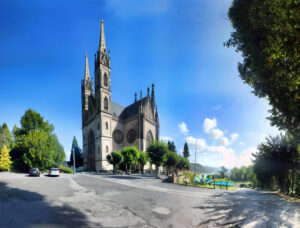 Panorama einer gotischen Kirche mit zwei hohen Spitztürmen vor blauem Himmel, gepflastertem Platz mit geparkten Autos und Bäumen sowie Blick auf See und Hügel.