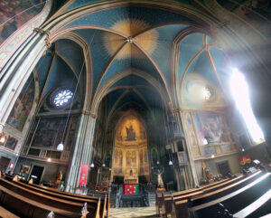 Innenansicht einer gotischen Kirche mit sternenverzierter Decke, bunten Wandmalereien und Sonnenstrahlen, die durch die Fenster strahlen.