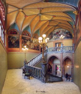 Prunkvoller Treppenaufgang mit kunstvollen Wandmalereien und Decke im historischen Gebäude. Beleuchtet von eleganten Kronleuchtern.