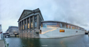 Panorama eines Stadtabschnitts am Wasser mit links einem klassizistischen Gebäude mit Säulen und Bögen und rechts einem modernen Glasbau, der über die Wasserfläche hinausragt.