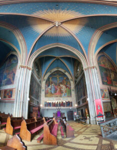 Innenansicht einer gotischen Kirche mit blauer, sternenverzierten Decke und bunten Wandmalereien. Bankreihen und ein thronartiger Stuhl im Vordergrund.
