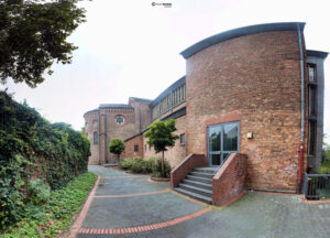 Außenansicht der modernen St. Augustinus Kirche in Bonn-Duisdorf aus roten Backsteinen mit grüner Bepflanzung und Gehweg.