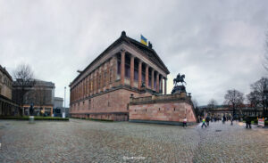 Großes klassizistisches Steingebäude mit ionischen Säulen und dreieckigem Giebel, davor ein Reiterstandbild auf gepflastertem Platz, umgeben von kahlen Bäumen und Menschen unter bedecktem Himmel.