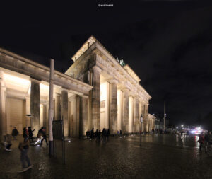 Brandenburger Tor bei Nacht, beleuchtet, mit zwei Passanten und angedeuteten Gebäuden im Hintergrund.