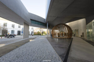 Moderner Außenbereich des MAXXI Museums in Rom mit einer einzigartigen Holzskulptur in Kugelform, umgeben von Betonwegen und Gebäuden bei sonnigem Wetter.