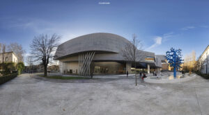Weitwinkelansicht des modernen MAXXI Museums in Rom mit seiner geschwungenen Betonfassade und abstrakten blauen Skulptur, umgeben von Bäumen und einem öffentlichen Platz unter blauem Himmel.