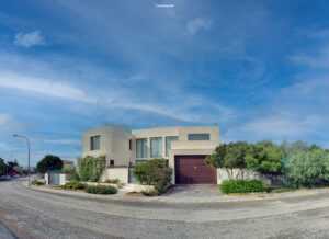Weitwinkelaufnahme einer modernen, hellen Villa mit Flachdach und großen Fenstern, umgeben von mediterraner Vegetation an einer kurvigen Straße unter blauem Himmel.