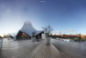 Zeitgenössisches Pavillon mit mehreren spitzen Zeltdächern auf hölzernem Deck, flankiert von großen Pflanzkübeln mit kahlen Bäumen und umgeben von urbanen Gebäuden unter klarem Himmel.