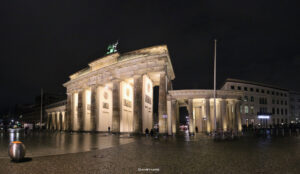Brandenburger Tor in Berlin bei Nacht, prachtvoll angestrahlt, mit zwei Passanten, Kopfsteinpflaster und angedeuteten Altbauten im Hintergrund.