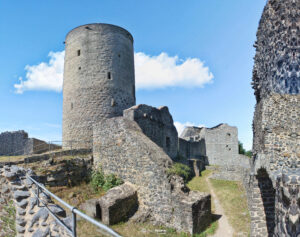 Ansicht der Nürburg Ruine in der Eifel mit massivem Rundturm, umgeben von weiteren Steinmauern und grünem Gras unter blauem Himmel mit Wolken.