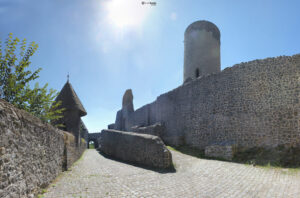 Kopfsteinpflasterweg innerhalb der Nürburg Ruine, flankiert von alten Steinmauern und einem markanten Rundturm, unter strahlendem Sonnenschein.