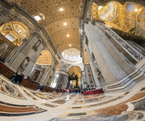 Weitwinkelaufnahme des Petersdoms in Rom, die den prunkvollen Innenraum mit den reich verzierten Decken, Säulen und den monumentalen Skulpturen zeigt.
