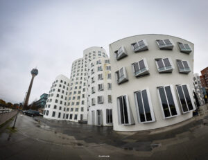 Moderne Wohn- und Bürogebäude mit weißer Fassade und schräg angeordneten Fenstern, nasses Pflaster im Vordergrund und hoher Fernsehturm vor bewölktem Himmel.