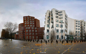 Zwei nebeneinanderstehende Gebäude – links ein rotes, modernes Backsteingebäude mit klaren Fenstern, rechts ein geschwungenes, glänzendes Metallbauwerk – vor nassem Kopfsteinpflaster und trüb bewölktem Himmel.