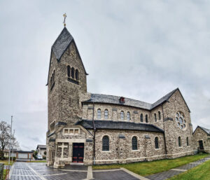 Steinerne Kirche mit hohem Glockenturm, Satteldach, Rundbogenfenstern, Doppelportal und Rosettenfenster, umgeben von gepflastertem Weg, Grasflächen und Bäumen unter bewölktem Himmel.