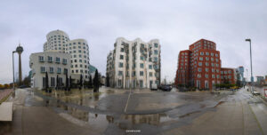Panorama einer modernen Stadtszene mit Fernsehturm, geschwungenen Fassaden, nassem Pflaster und Regenschirmen an Außenbestuhlung.