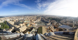 Panoramablick von oben auf den Petersplatz, die Vatikanstadt und das historische Rom mit seinen Gebäuden und dem weitläufigen Stadtbild unter blauem Himmel.